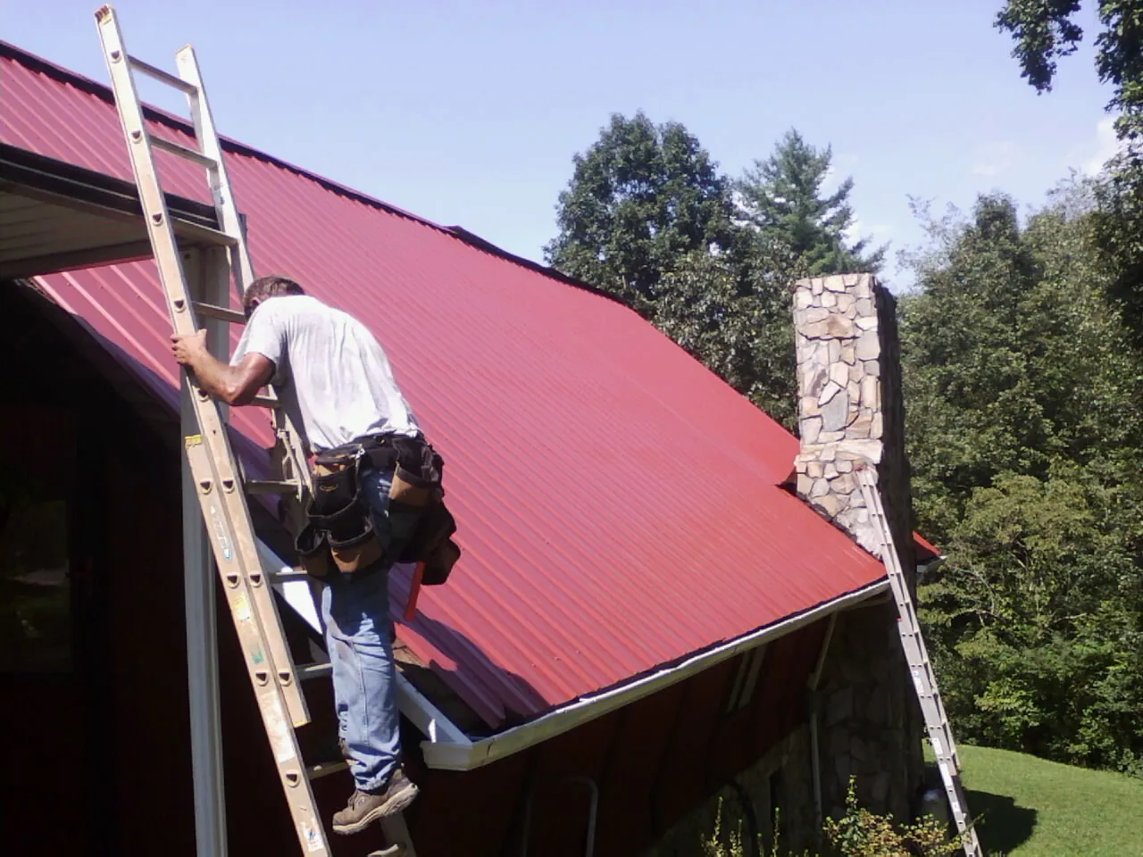 Roofer on a ladder beside a red metal roof and stone chimney — Soffit & Fascia Repair by Roofing Brothers in Mayer